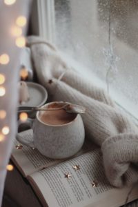 A cup of hot cocoa sitting on an opened book that has little plastic golden stars on it. The book is lying on a window ledge nestled next to a cozy, white blanket. There is snow and ice on the outside of the window. 