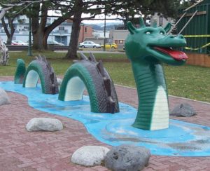 An Ogopogo statue in British Columbia. It is green and grey and has parts of its body poking up from a blue stream. 