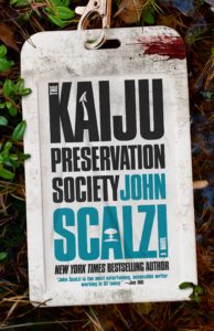 The Kaiju Preservation Society by John Scalzi Book cover. Image on cover shows the title and author of the book written on a blood-covered tag that’ surrounded by green leaves. 