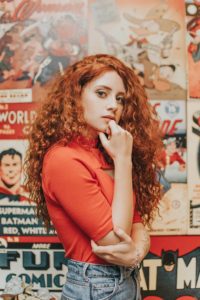 A red headed woman with long, curly hair standing in front of a wall plastered over with magazine covers. 