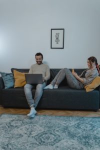 A man using a laptop while sitting on a couch. A woman has stretched out next to him on the cough and has her legs by his thighs as she listens to something. 