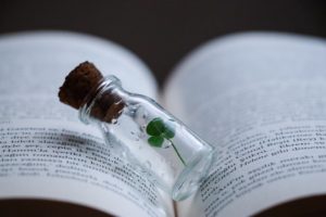 Three leaf clover in a little glass jar with a cork stopper. The jar is sitting in the crease of an opened book. 