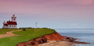 A lighthouse on Prince Edward Island. You can see the famous red sandy beaches between the lighthouse and the sea. The sky is a dusky rose colour. It may be dusk or dawn in this photo. 