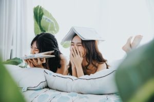 Two women laughing while reading books. One has a book on top of her head. 