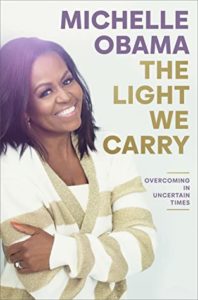 The Light We Carry by Michelle Obama book cover. Image on cover is a photo of her smiling, crossing her arms in a hug, and wearing a white and tan sweater. 