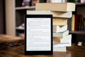 An ereader propped up by eight paper books that are stacked on top of each other. 