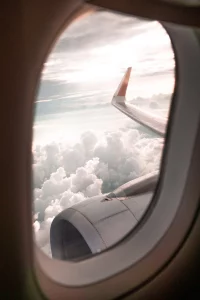 Photo taken peering out of a windows on a plane. You can see one engine, part of a wing, and lots of fluffy white clouds. 