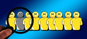 A row of yellow smiley face people. There is one grey person among them who isn’t smiling, and the magnifying glass is focused on that person. 