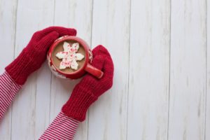 Person wearing red mittens and a a red and white striped shirt holding a red mug filled with hot cocoa and a white star cookie sprinkled with red sprinkles that is sitting in the cocoa. 