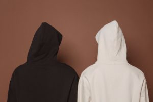 Two people wearing hoodies and facing a brown wall. One person has a black hoodie and one has a white hoodie. 