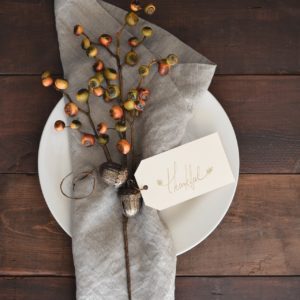 A white plate covered with a grey napkin that has a sprig of red and brown berries and a card that says “thankful.”