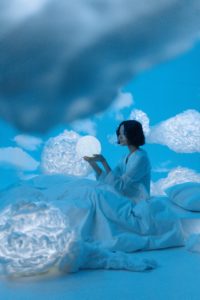An Asian woman sitting upright in a white bed. The walls are painted to look like a semi-cloudy blue sky, and there’s a big, dark cloud right in front of her. She’s holding a glowing white orb in her hands. 