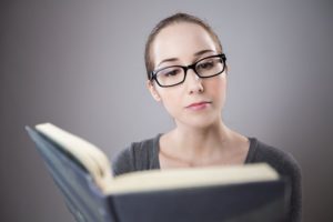 White woman wearing black-rimmed glasses and looking studious as she reads a hardback book. 