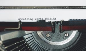 A typewriter with a white sheet of paper stuck in it. The phrase “something worth reading” has been typed on to the sheet of paper. 