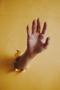 A white person’s hand and forearm has punched through a yellow wall and is reaching out for help with all five fingers extended. 
