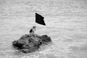 Black and white photo of a white woman wearing a black one-piece bathing suit. She’s crouching on a barren rock that’s surrounded by water and placing a black flag on the rock. The waves around her are gentle, and it appears to be a nice day outside although the sky is not visible. 