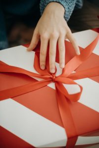 Close-up of the hand of a white person as they begin unwrapping a red ribbon that’s been tied into a bow around a box wrapped in red and white striped paper. 