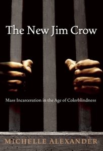 Book cover for The New Jim Crow: Mass Incarceration in the Age of Colorblindness by Michelle Alexander. Image on cover shows the hands of a brown-skinnned person clutching the bars of a cell as they stand in the darkness of the cell all around them. 