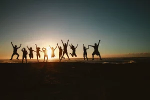 Ten people standing on top of a hill just as the sun has slipped below the horizon and darkness covers three quarters of the land. You can see the silhouettes of their bodies as they all leap for joy with their knees bent and their arms outstretched above them. 