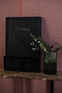 The word goodbye is written in white chalk on a black chalkboard. The chalkboard is sitting in a black mesh container on a wooden shelf. There is a plant growing out of a green drinking glass next to the chalkboard. 