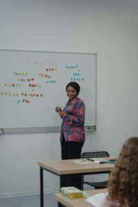 A black female teacher wearing black pants and a floral pink, purple, blue, and red blouse standing in front of a whiteboard. She has written the word English on the board and is looking at her students as they give her other words to put below it. Names like “Ringa,” “EBJ,” “Klas” have already been placed there using plastic alphabet letters that are in bright primary colours like red, blue, and green. 