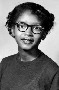 A black and white photograph of Claudette Colvin in 1952, three years before she refused to give up her seat to a white person. She is wearing thick black glasses in this photo and a dark grey shirt. She is smiling faintly at the camera with her head turned on a slight angle. Her hair is neatly pulled behind her head. 