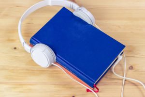 A white pair of headphones that have had their two ear pieces placed on either side of a blue hardback book. The plug in the headphones has been tucked between the book’s pages as if to magically absorb their words and turn them into an audiobook. It made me chuckle. 
