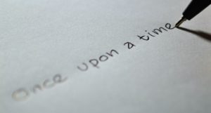 A closeup of a black ballpoint pen that has just finished writing the phrase “once upon a time” in black ink on a white unlined piece of paper. 