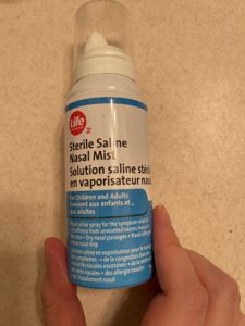 The small hand of a Caucasian adult is holding a blue and white bottle of Life Brand Sterile Saline Nasal Mist. 