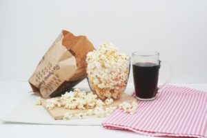 A clear glass bowl filled with popcorn is sitting on a white table. There is a clear glass filled with a dark soda sitting on a red and white checkered napkin on the left hand side of the popcorn bowl. On the right hand side, an opened bag of microwave popcorn is leaning on the glass bowl of popcorn and there are about two dozen pieces of popcorn spilled over the white table and red and white checkered napkin. 