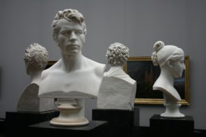 Four busts in a museum. Three are men and one is a woman. Three have curly hair and the woman has her straight hair in a pony tail. Each bust is facing a different way, so only the faces of one man and one woman can be seen. 