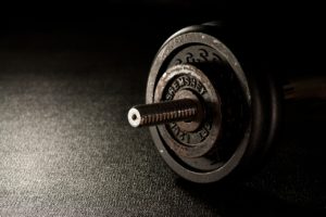 A closeup of a dumbbell sitting on a grey flat floor. Sunlight is streaming into the room and just barely reaching the end of the dumbbell. 