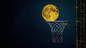 A photograph of a basketball net against a dark night sky. The photo is positioned so that it looks like the full moon in the sky is about to swish through the basketball net. 