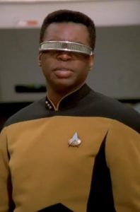Photo of LeVar Burton playing Geordi La Forge on Star Trek: The Next Generation. He is wearing a yellow Star Trek uniform and his visor and looking ahead of himself with a serious expression on his face. 