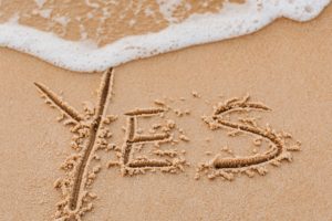 The word yes is written into sand on a beach. A wave is just beginning to reach the top of the letter y in yes and erase it. 