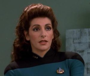 Photo of Marina Sirtis playing Deanna Troi on Star Trek:Deep Space Nine. She is wearing a teal Star Trek uniform and looking ahead of her seriously. 