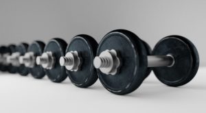 A row of black dumbbells lined up neatly and orderly on a white floor. The wall behind them is white as well. 