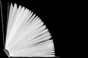 Black and white photo of a hardcover book that is opening. All of it’s white pages are gently fanning out as the cover slowly moves down towards the the surface the book is lying on. 