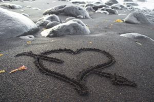 A black and white photo of a heart drawn into sand on a rocky beach. There is an arrow drawn through the heart and several white rocks that are about the size of a human infant sitting in the sand behind the drawing. 