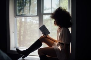 A black woman with a large Afro is sitting on the ledge of a window in a dark room. Light is pouring into the room around her as she holds up a hardback book to the light and reads. 