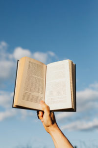 The arm of a person with pale skin is holding an opened hardcover book up against a blue sky that has a few fluffy white clouds sailing through it. 