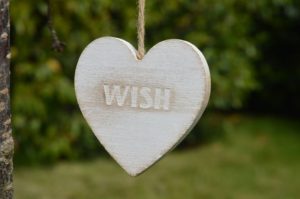 A white wooden heart with the word “wish” carved into it. The heart is tied to something out of range of the shot with a piece of twine. There is a blurry, out-of-focus forest behind the heart and green grass below it. 