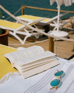 There are three yellow lounges on a wooden deck by the sea or a large lake. The lounge closest to the viewer also has a white and grey beach towel, a pair of sunglasses, and an opened hardback book on it. 