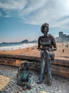 A metal statue of a woman sitting on a stone ledge. She’s holding a book and looking up at the land. There is a metal statue of a dog looking in the same direction she is. The dog is sitting on it’s haunches but has it’s front legs touching the land as if it’s about to stand up fully and run away. 