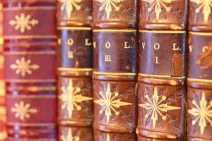Closeup photo of hardback, antique books with red covers and gold stars on their spines. The words “Volume I,” “Volume II,” and “Volume III” are stamped in gold ink on their spines. 