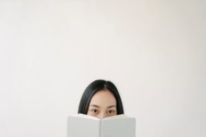 An Asian woman smiling slightly as she holds a hardback book over the lower half of her face and peeks out above it at the viewer. The book has a white cover with no title or author printed on it, and the woman is standing in a similarly white and barren room. 