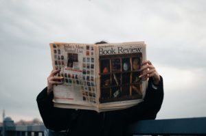 A white person wearing a black sweatshirt is holding up the Book Review section of the New York Times while standing outside on a cloudy day.