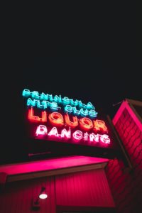 A neon sign lit up against a black night sky in a city. You can see a skyscraper next to the sign. The words “Peninsula Night Club” are in neon blue on the sign. The word “liquor” is larger than all other words and in neon orange on the sign. The word “dancing” is on the bottom of the sign and in neon pink. 