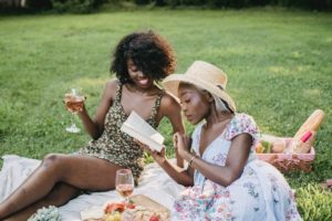 Two pretty black women are wearing sundresses and having a picnic on a blanket in the park. The one on the left is wearing a straw hat and reading a book to her companion while the other one sips a glass of wine and smiles. 