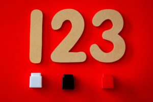 The numbers 1, 2, and 3 are beige coloured and lying on a red surface. Beneath each letter is a small lego: a white one underneath the number 1, a black one underneath the number 2, and a red one underneath the number 3. 
